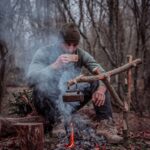 An adult man enjoys coffee by a campfire in a serene forest setting.