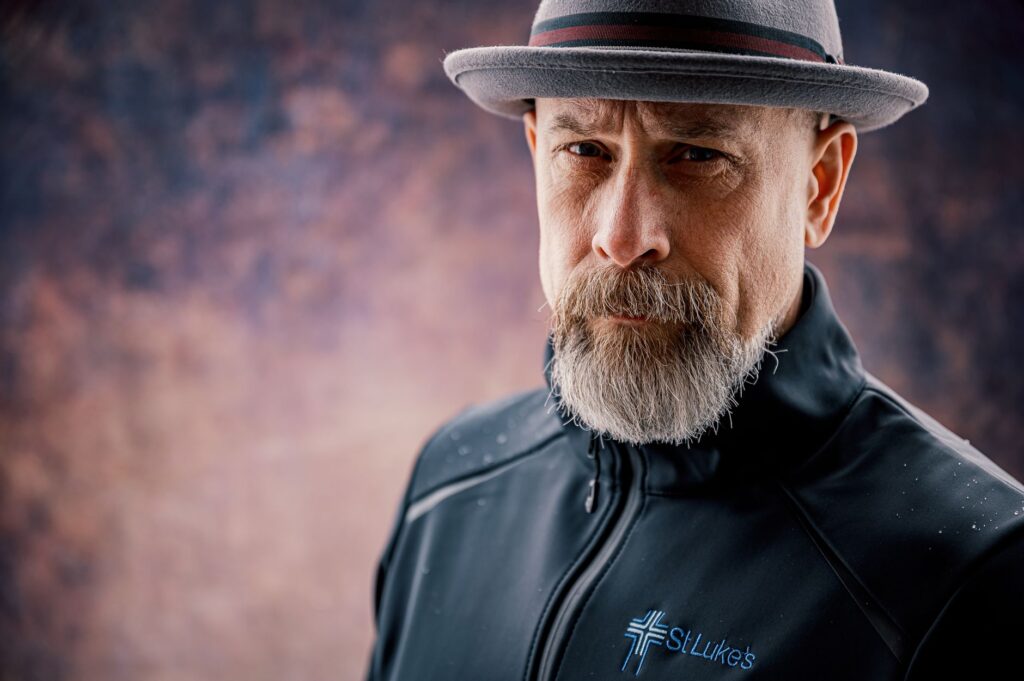 Portrait of a mature bearded man wearing a fedora and a jacket with a serious expression.