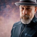 Portrait of a mature bearded man wearing a fedora and a jacket with a serious expression.