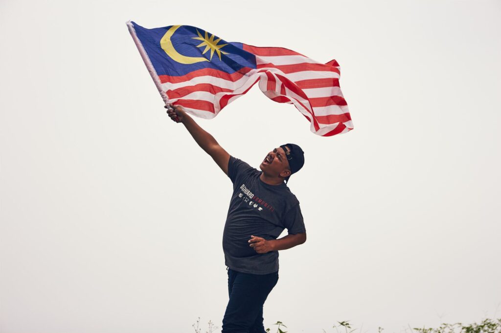 A man proudly waves the Malaysian flag outdoors, expressing joy and patriotism.