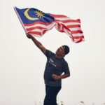 A man proudly waves the Malaysian flag outdoors, expressing joy and patriotism.
