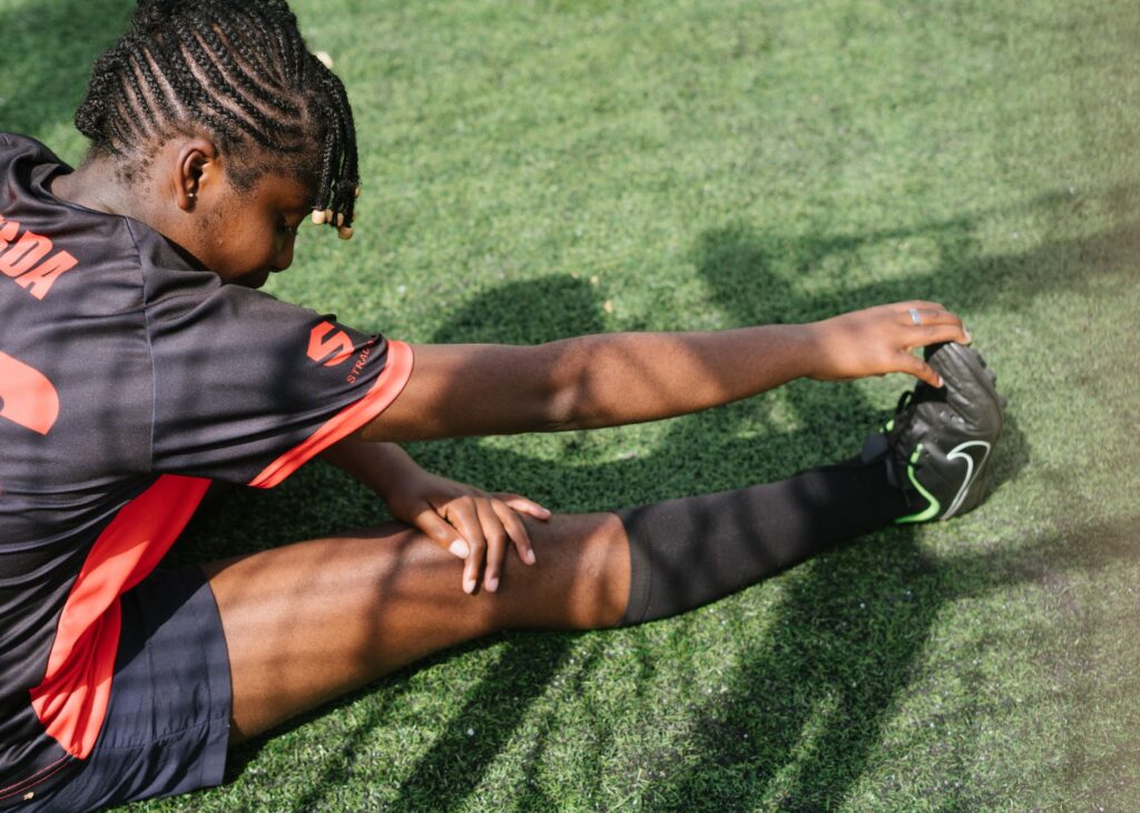 From above of black female soccer player sitting on green grass and stretching before coming on field