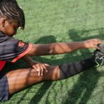 From above of black female soccer player sitting on green grass and stretching before coming on field