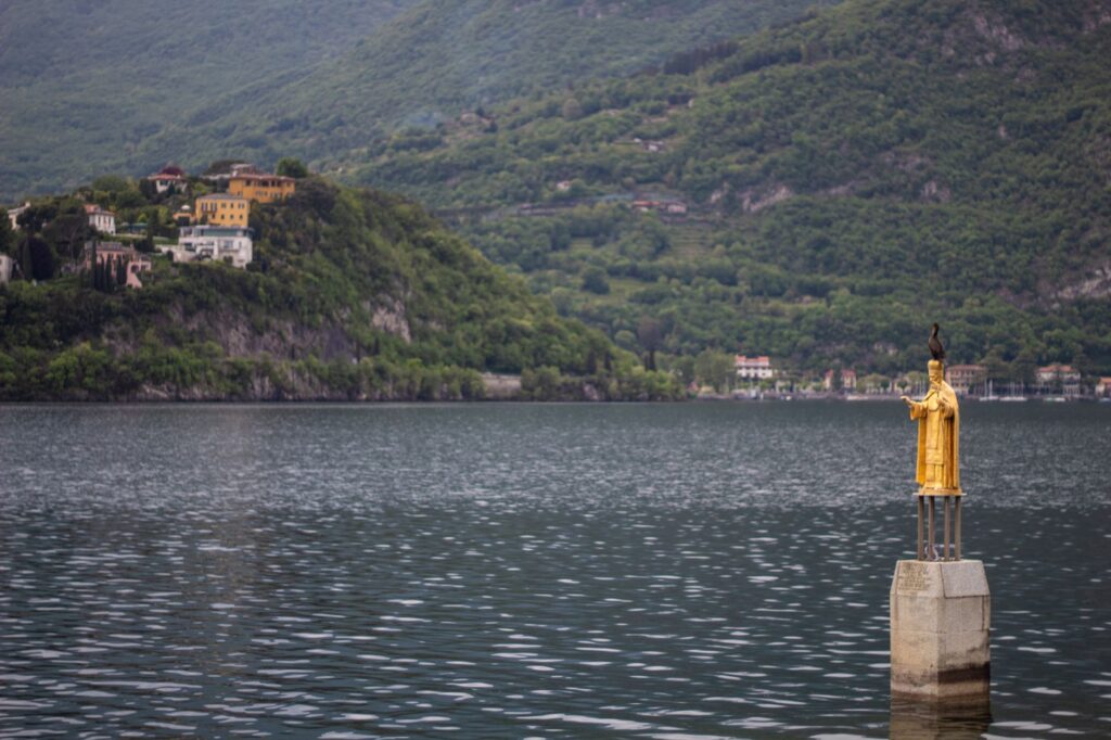A serene view of a golden statue on Lake Como with lush greenery in the background.