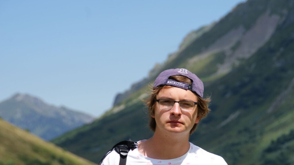 Young man hiking in the mountains, wearing a cap and glasses, enjoying the summer outdoors.