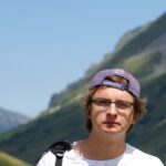 Young man hiking in the mountains, wearing a cap and glasses, enjoying the summer outdoors.
