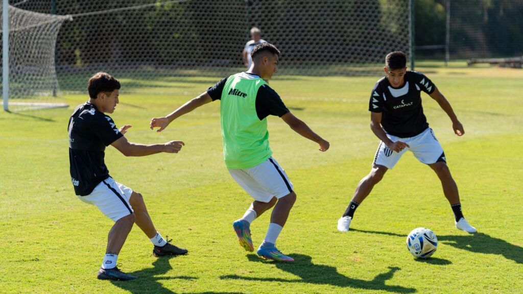 Three young men engage in soccer practice outdoors on a sunny day.