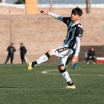 Teenage soccer player kicking a ball during a match on an outdoor field.