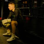 A male boxer sits with gloves on, contemplating in a dimly lit boxing ring.
