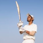 A cricket player in white uniform holds a bat, ready to play under the clear blue sky.