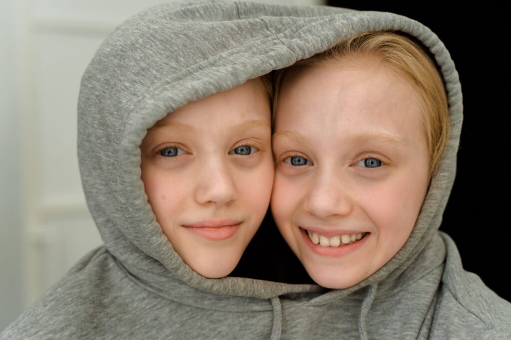 Charming close-up of smiling twin girls with blonde hair sharing a grey hoodie indoors.