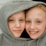 Charming close-up of smiling twin girls with blonde hair sharing a grey hoodie indoors.