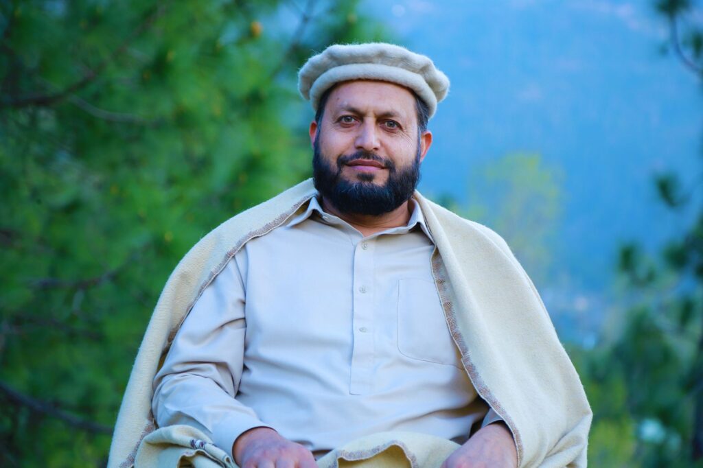 A South Asian man wearing a Chitrali cap and shawl, seated outdoors with a serene backdrop.