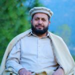 A South Asian man wearing a Chitrali cap and shawl, seated outdoors with a serene backdrop.