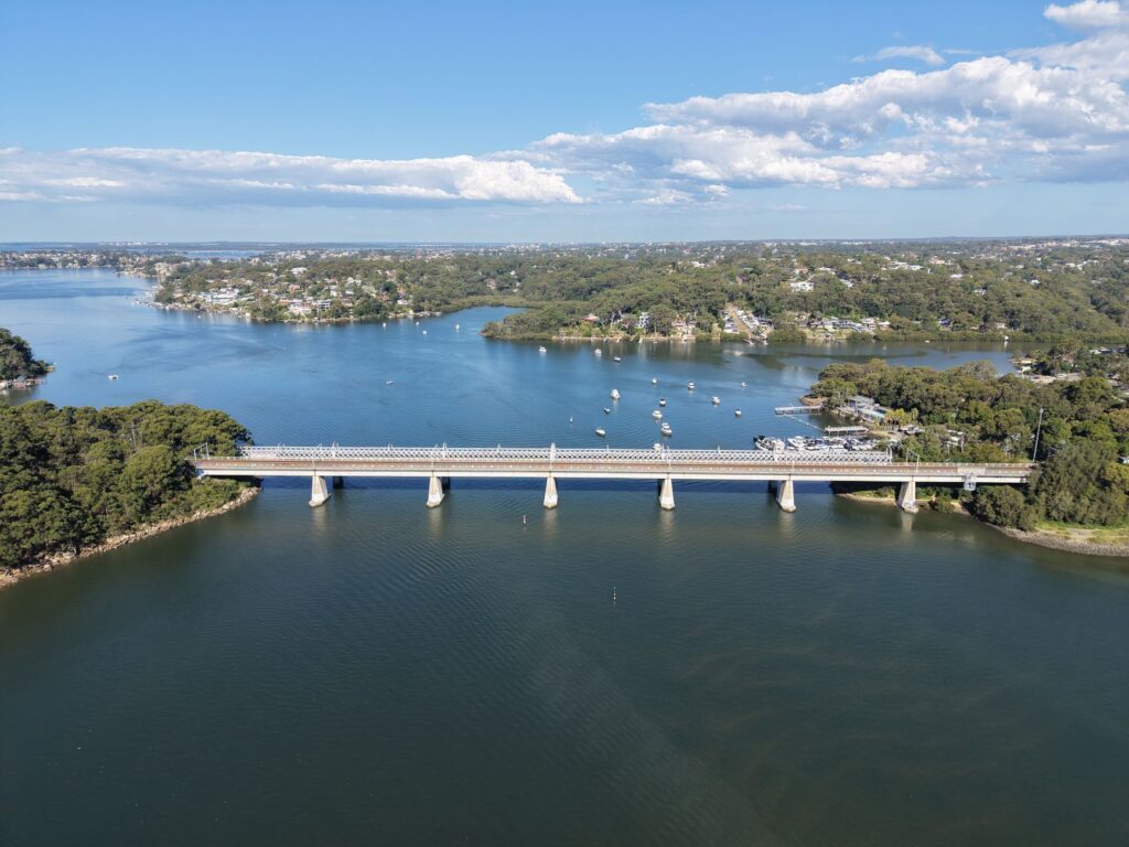 Aerial view of Captain Cook Bridge spanning over Georges River, connecting lush landscapes under a clear blue sky.