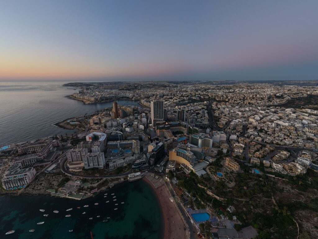 Discover a breathtaking aerial view of St. Julian’s, Malta as the sun sets, illuminating the coastline and cityscape.