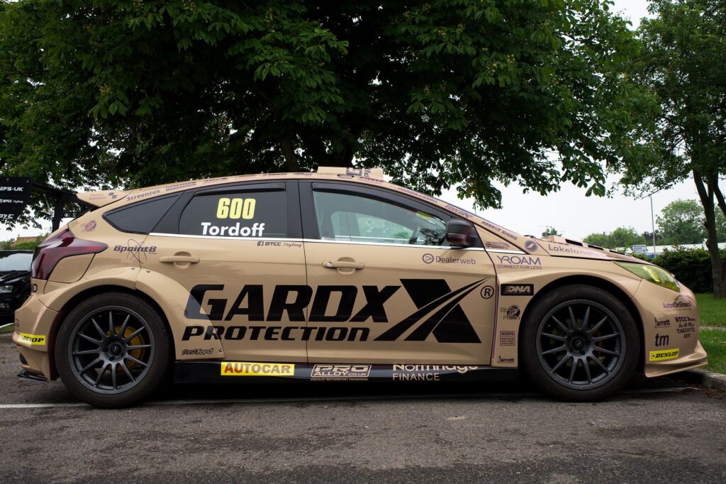 A side view of a racing car with promotional decals, parked under a green tree.