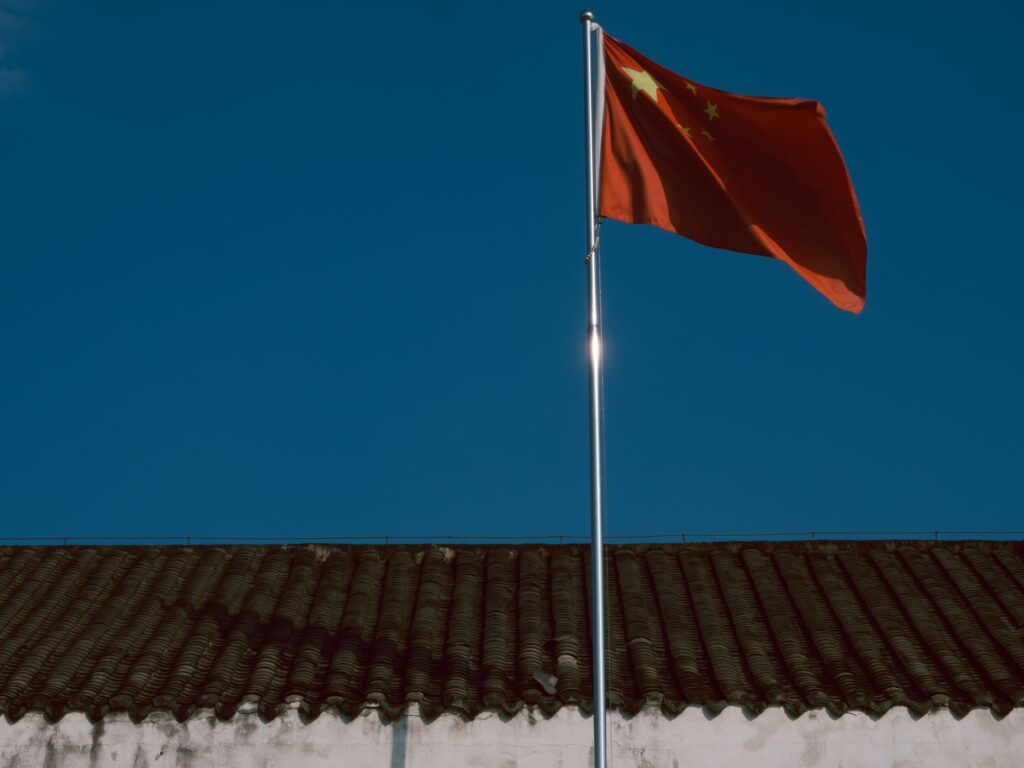 Chinese flag waving over traditional building roof against a clear blue sky.
