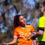 Female soccer player in orange jersey argues with referee on the outdoor field during a sunny day match.