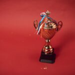 Golden trophy cup with red, blue, and white ribbons on a red backdrop symbolizes achievement and success.