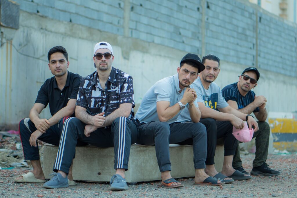 Five young men in urban casual attire posing outdoors against a concrete wall.