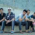 Five young men in urban casual attire posing outdoors against a concrete wall.