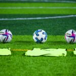 Three soccer balls and training bibs on a lush green field in Rawalpindi, Pakistan.