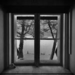 Black and white photo capturing a tranquil snow-covered landscape through a window, invoking a peaceful winter atmosphere.