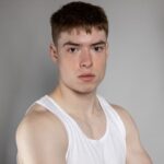 Portrait of a young man with freckles wearing a white tank top against a gray background.