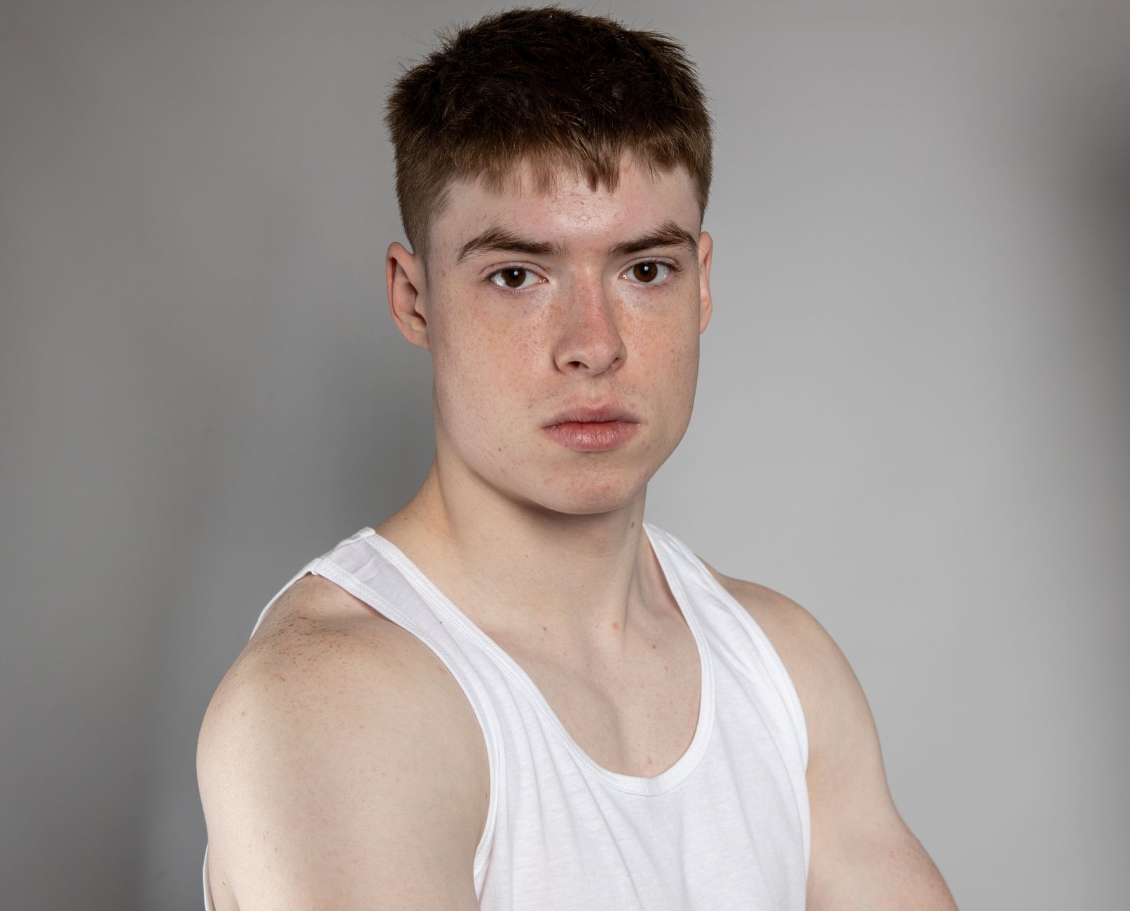 Portrait of a young man with freckles wearing a white tank top against a gray background.