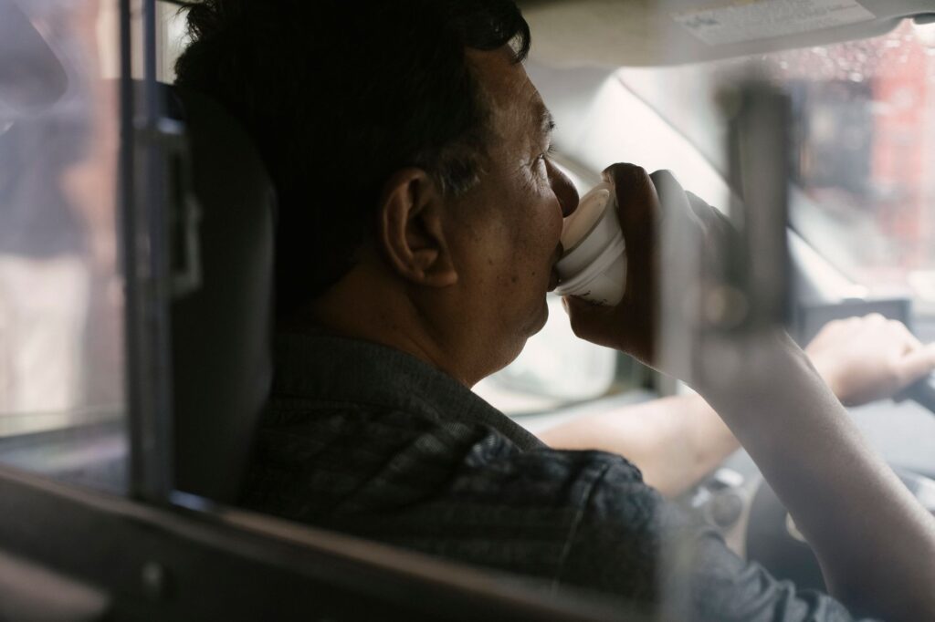 Back view of ethnic man drinking coffee in car while working as driver
