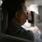 Back view of ethnic man drinking coffee in car while working as driver