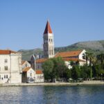 Scenic view of Trogir waterfront with the bell tower of St. Lawrence Cathedral against a mountainous backdrop.