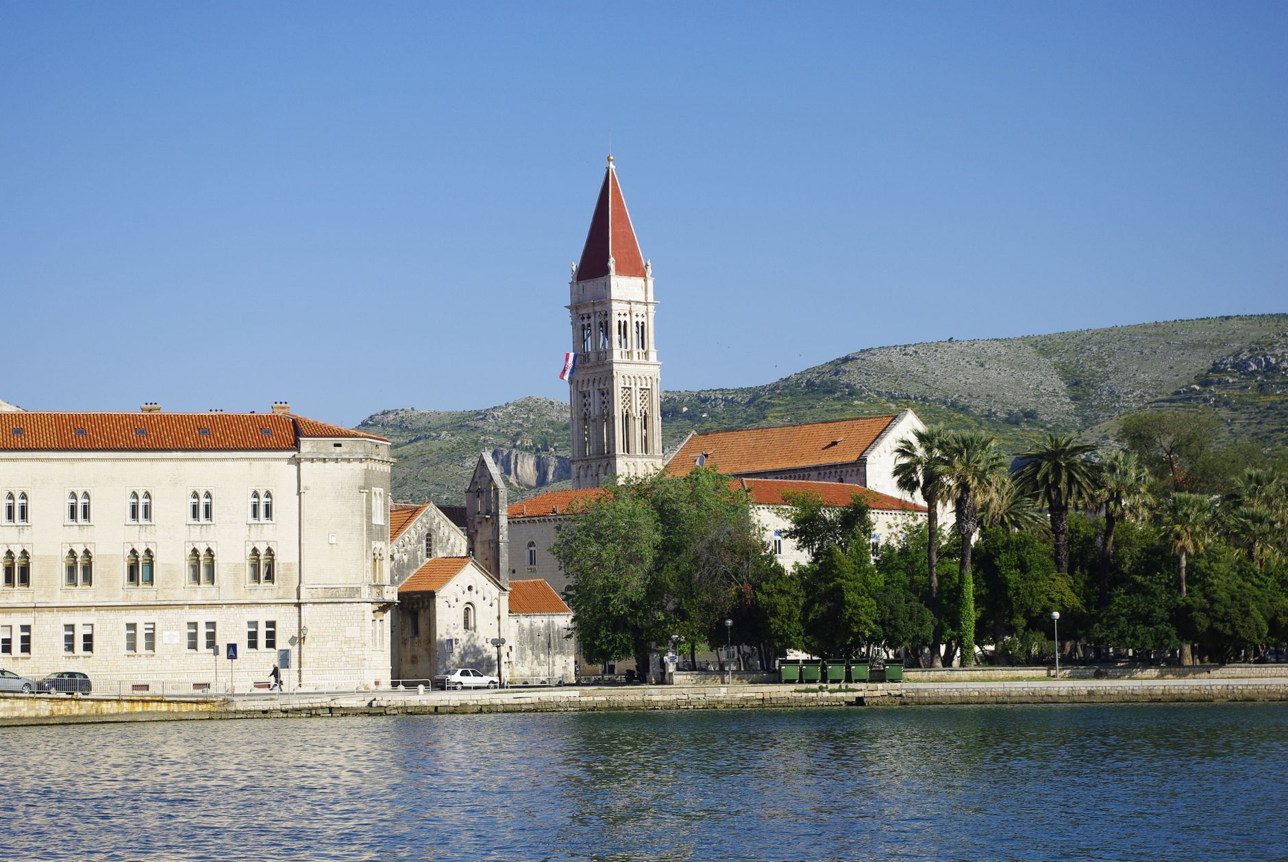 Scenic view of Trogir waterfront with the bell tower of St. Lawrence Cathedral against a mountainous backdrop.