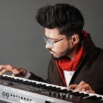 Man with glasses and red scarf playing an electronic piano intently.