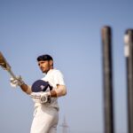 A cricket player in uniform poses with a bat on an outdoor field, ready for action.