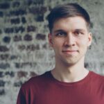 Casual portrait of a young man in a red shirt standing against a brick wall.