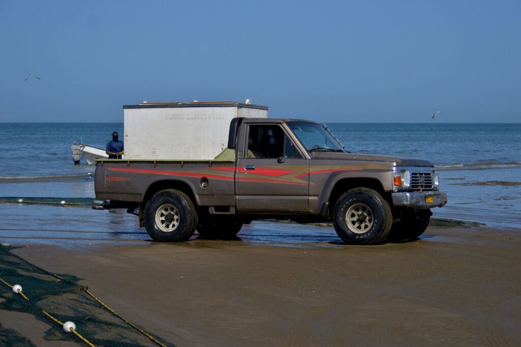 A brown pickup truck on a sandy beach with fishing nets and the ocean in the background.