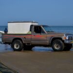 A brown pickup truck on a sandy beach with fishing nets and the ocean in the background.