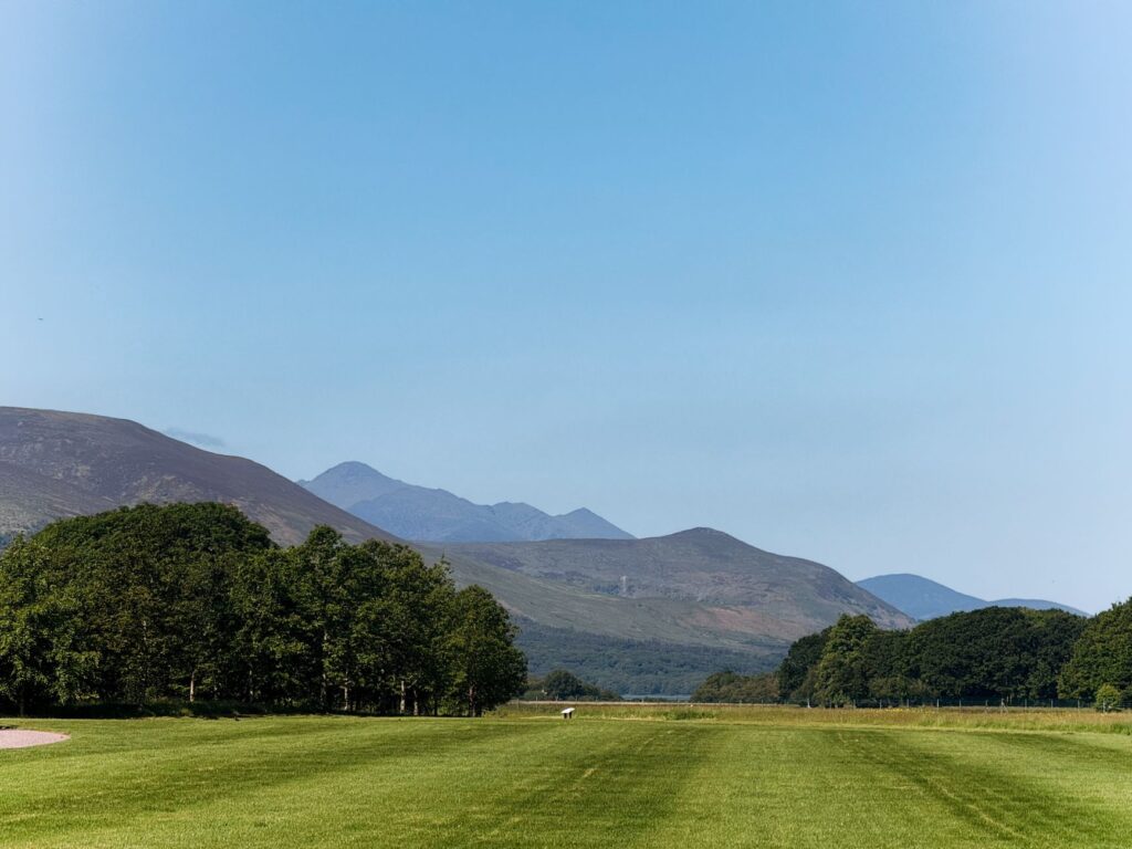 A serene view of the lush green fields and mountains of Killarney, Ireland under a clear blue sky.