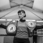 A female boxer confidently poses with championship belts in a boxing ring, showcasing strength and achievement.
