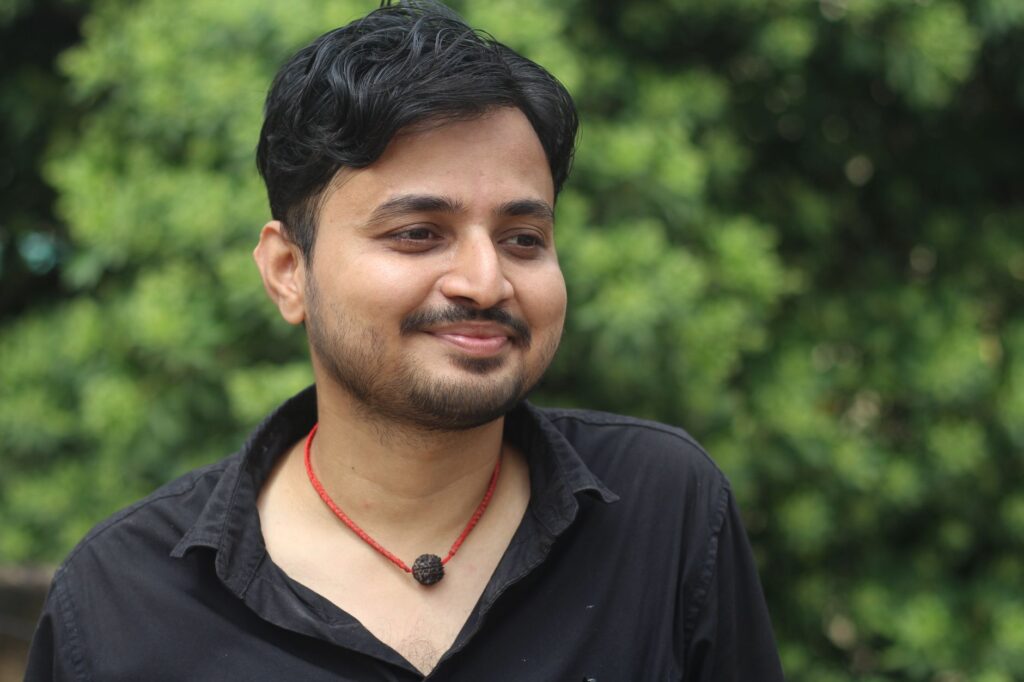 Portrait of a young man with a beard smiling outdoors against a green background in India.