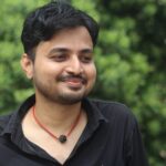 Portrait of a young man with a beard smiling outdoors against a green background in India.