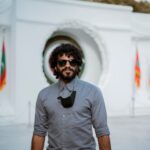 A man with curly hair and sunglasses poses in front of a monument with Maldives flags.