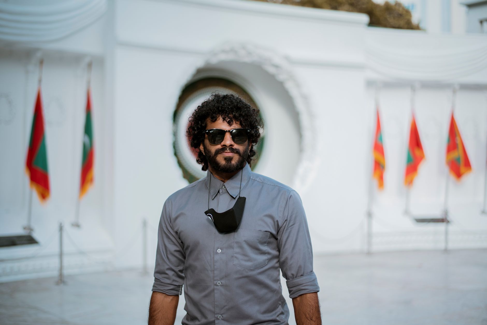 A man with curly hair and sunglasses poses in front of a monument with Maldives flags.