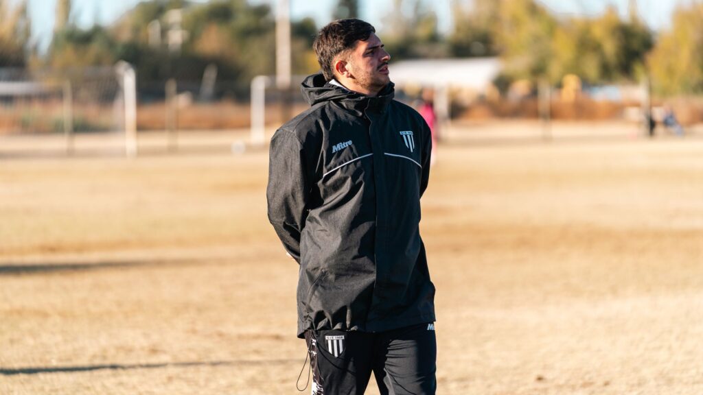 A soccer coach in black attire observes a training session on an outdoor field during daytime.