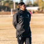 A soccer coach in black attire observes a training session on an outdoor field during daytime.