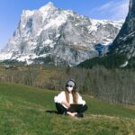 A woman sits with a laptop wearing a mask in a scenic mountain landscape, embracing remote work outdoors.
