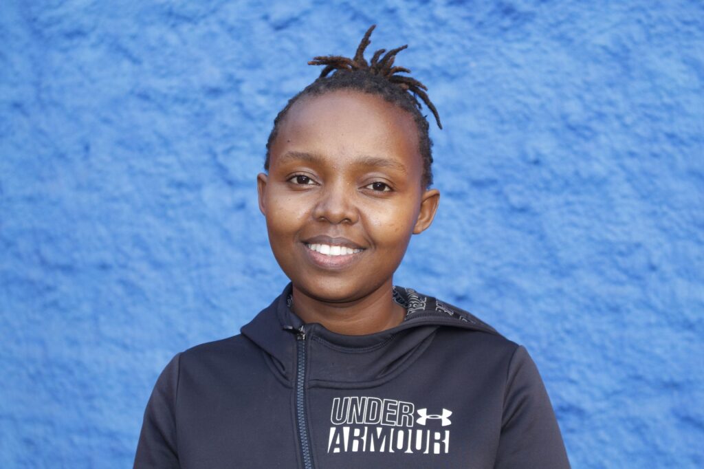 Portrait of a cheerful woman in a black jacket standing before a vibrant blue wall.