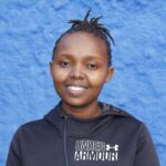Portrait of a cheerful woman in a black jacket standing before a vibrant blue wall.